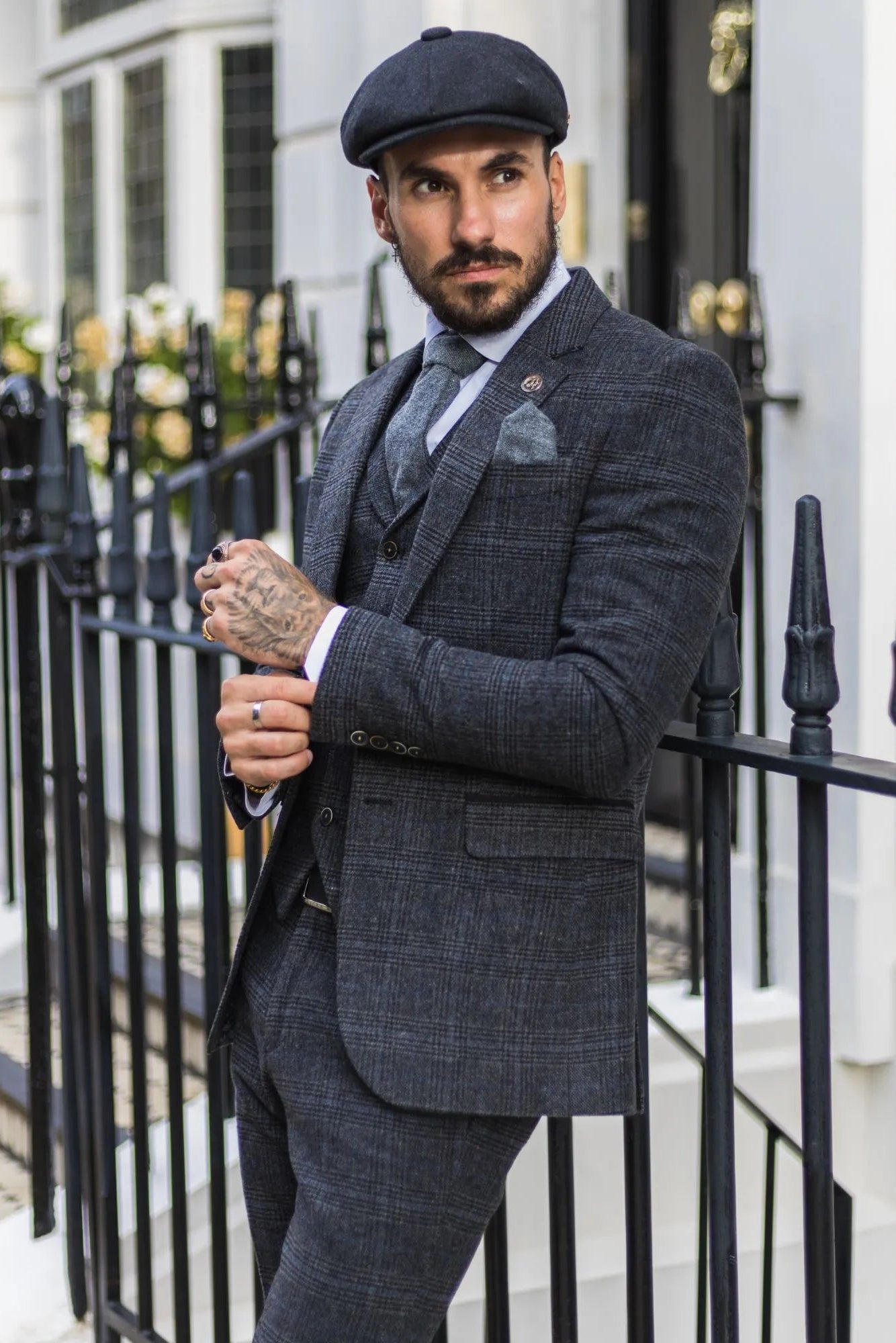 Man in a gray checkered suit standing outdoors next to a black metal gate.